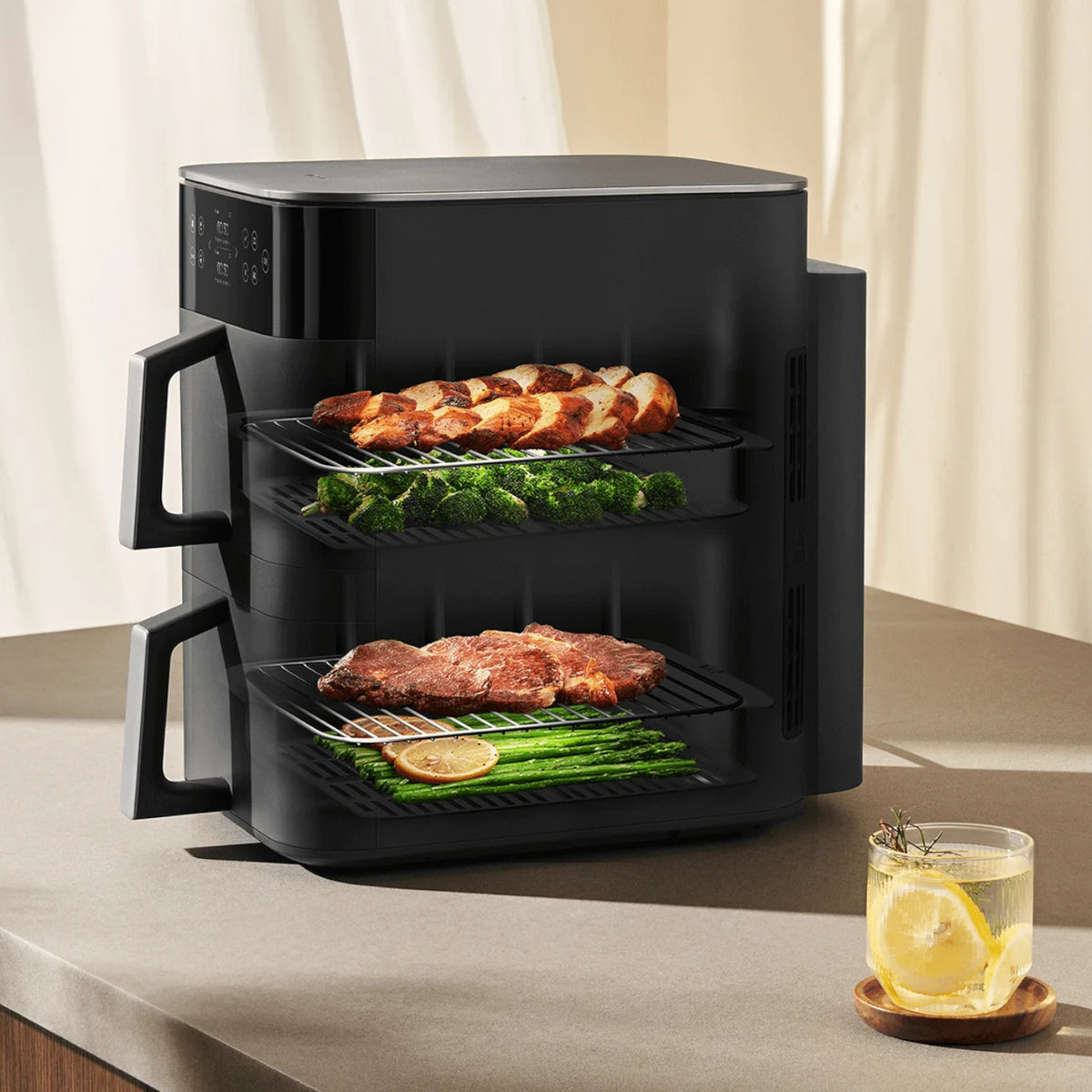Black air fryer with food on a countertop next to a glass of lemon water.