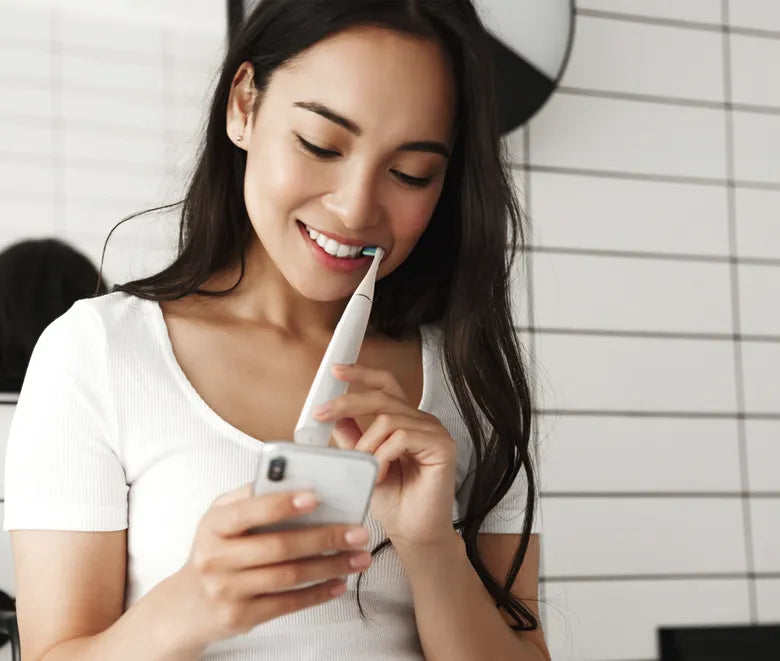Woman using a smartphone with a Aeno DB1S toothbrush in her mouth, against a white tiled wall.