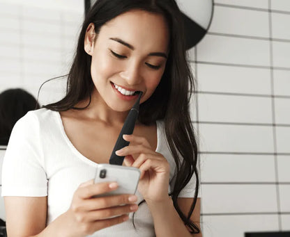 Woman using a smartphone with a Aeno DS2S Black Sonic Toothbrush in her mouth against a white tiled wall.