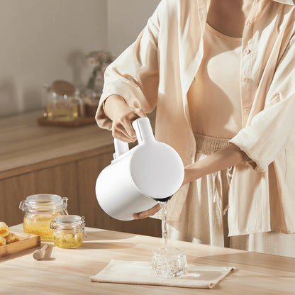 Person pouring water from a white teapot into a glass on a wooden table.