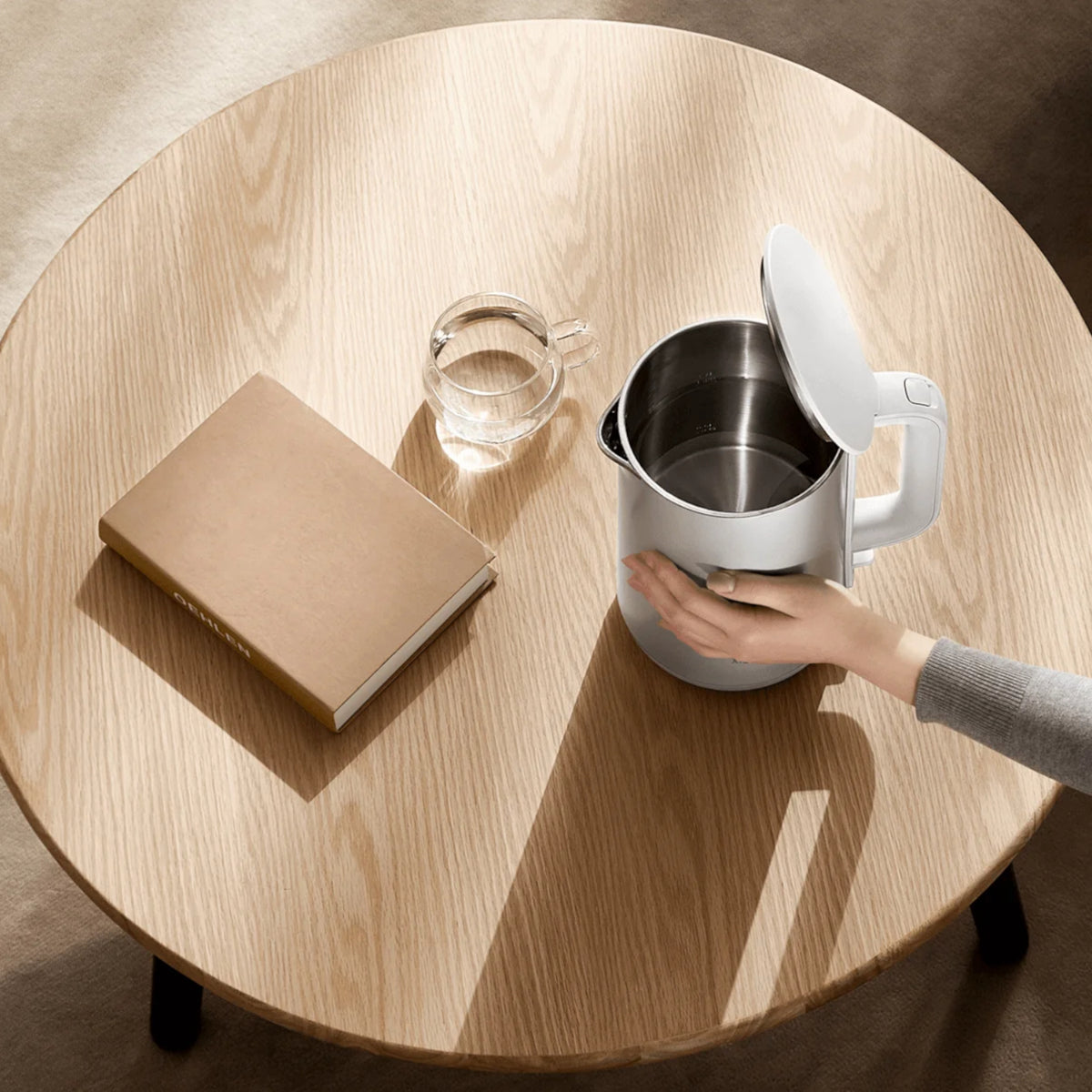 Person pouring water from a white electric kettle into a glass on a wooden table.